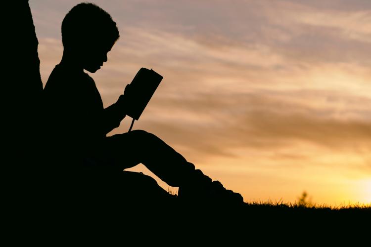 Silhouette of child reading a book outdoors. Photo by Aaron Burden, Unsplash. 