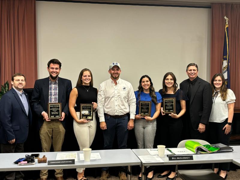 Several people pose for a photo in a conference room setting. Four young adults are holding plaques. 