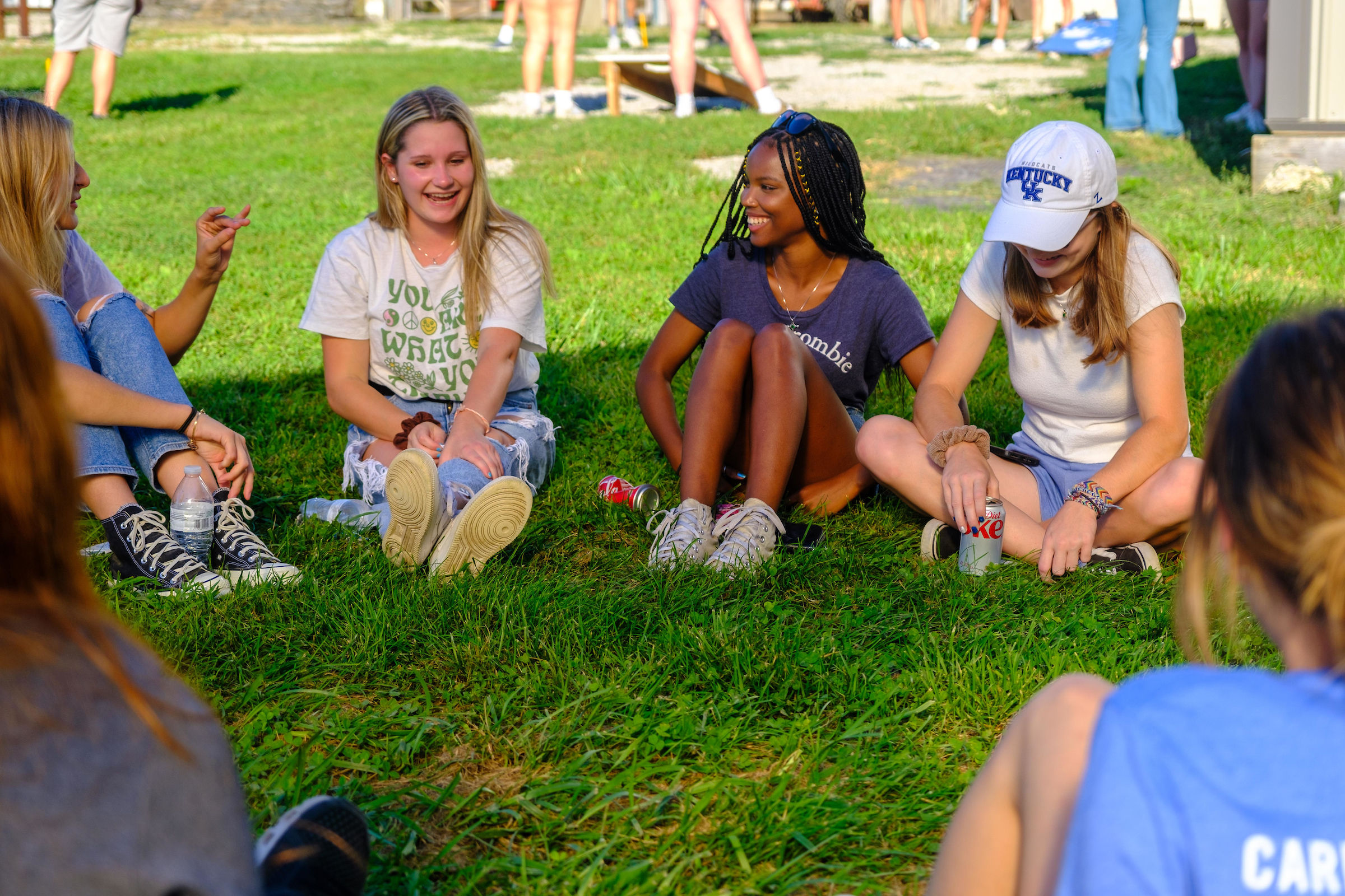 A group of young women sit in a circle on the grass, talking and laughing.