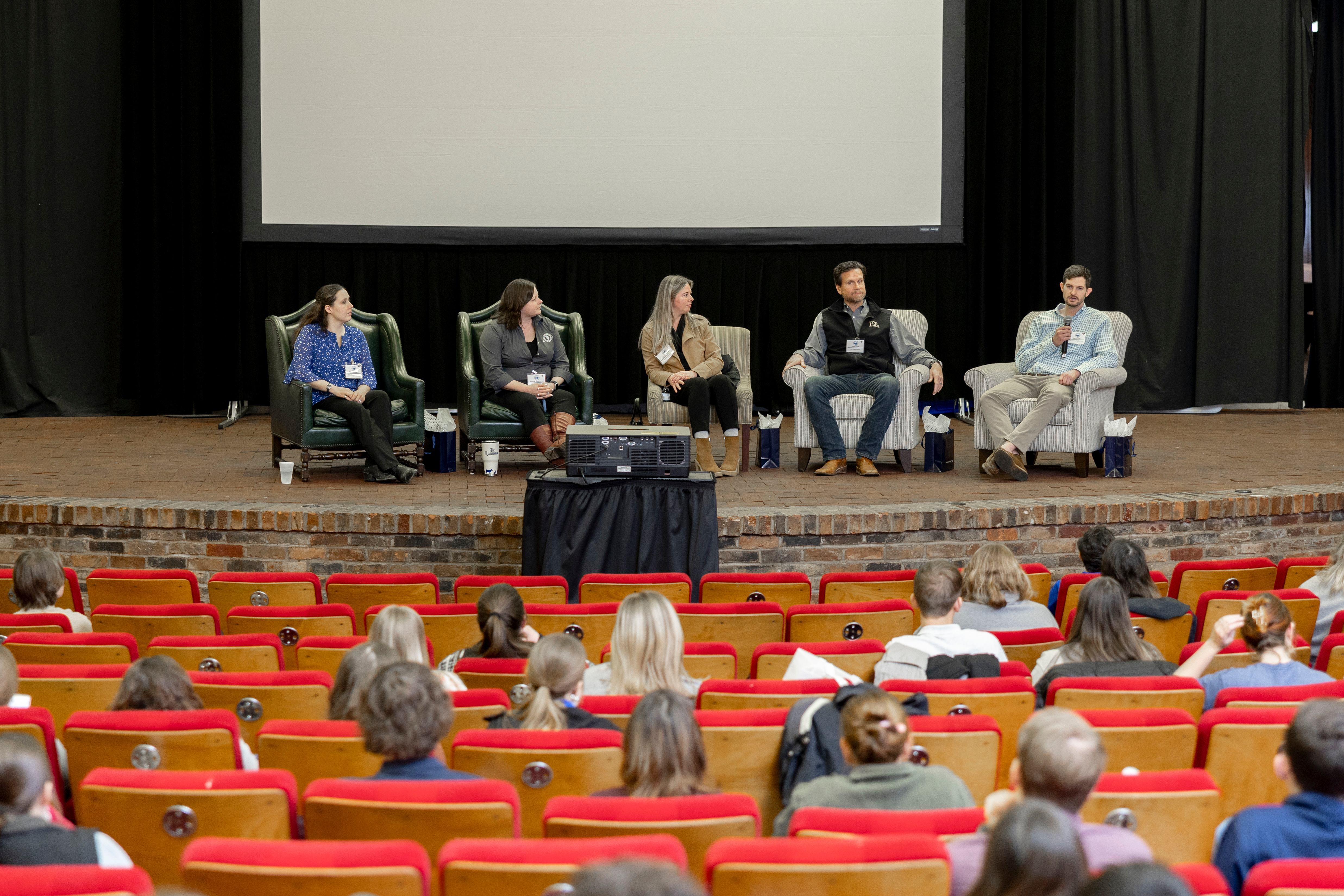 In a theater, people sit in the red seats and listen to a group of people speaking on stage.