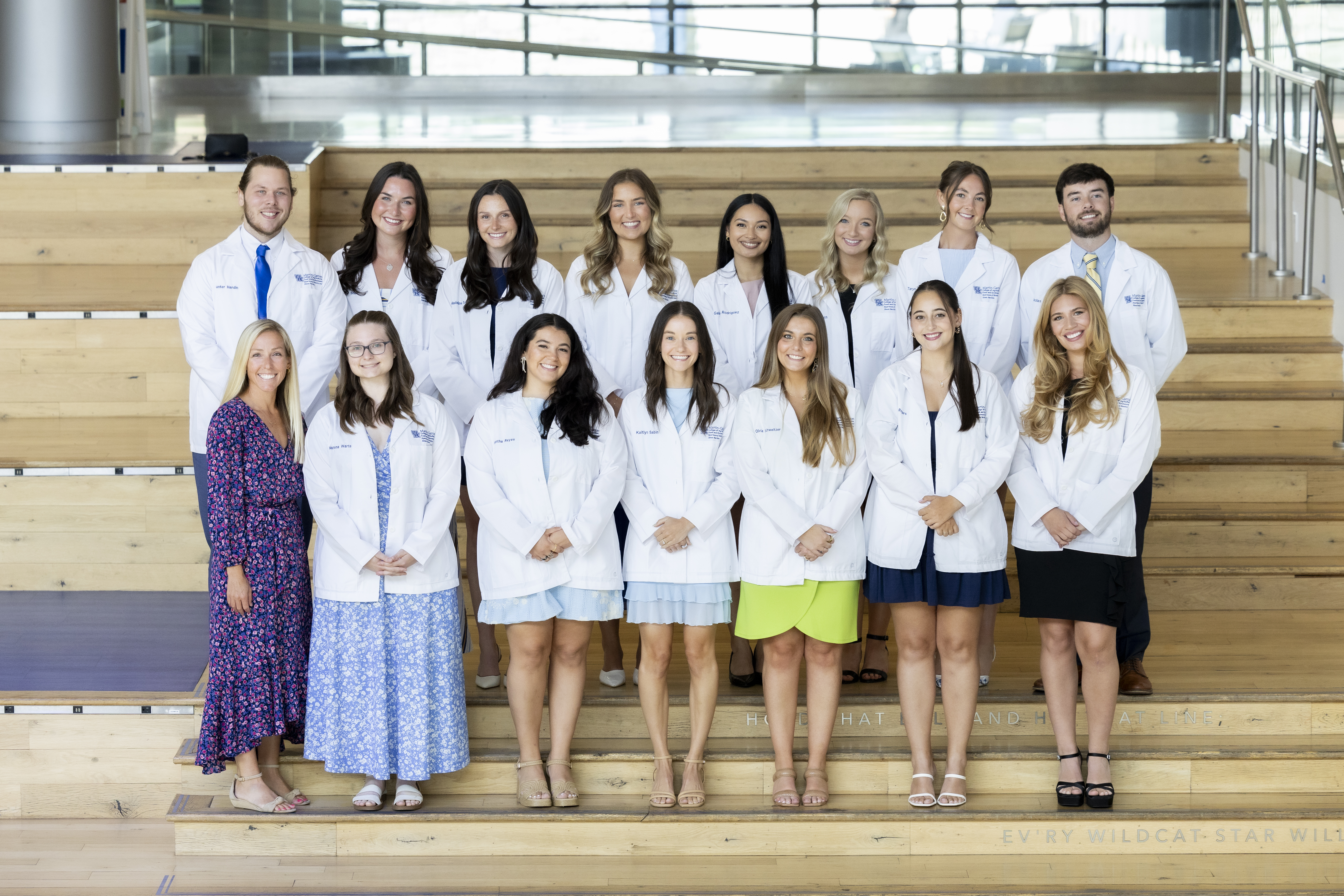 A group of 14 students wearing white dietitians coats and one woman in a purple dress pose for a photo on an internal staircase.