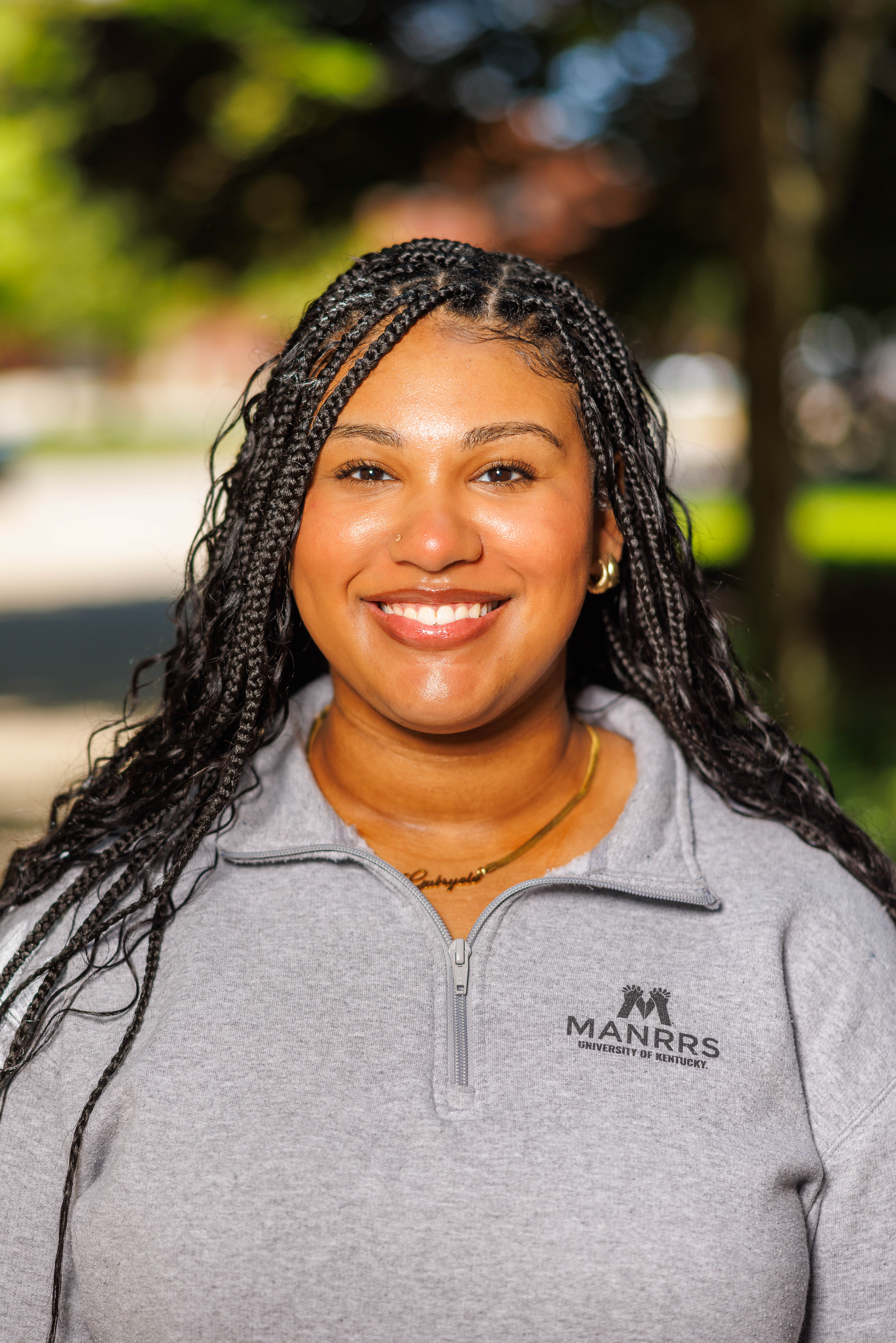 A young Black woman poses for a portrait on a college campus. Her pullover has the MANRRS logo.