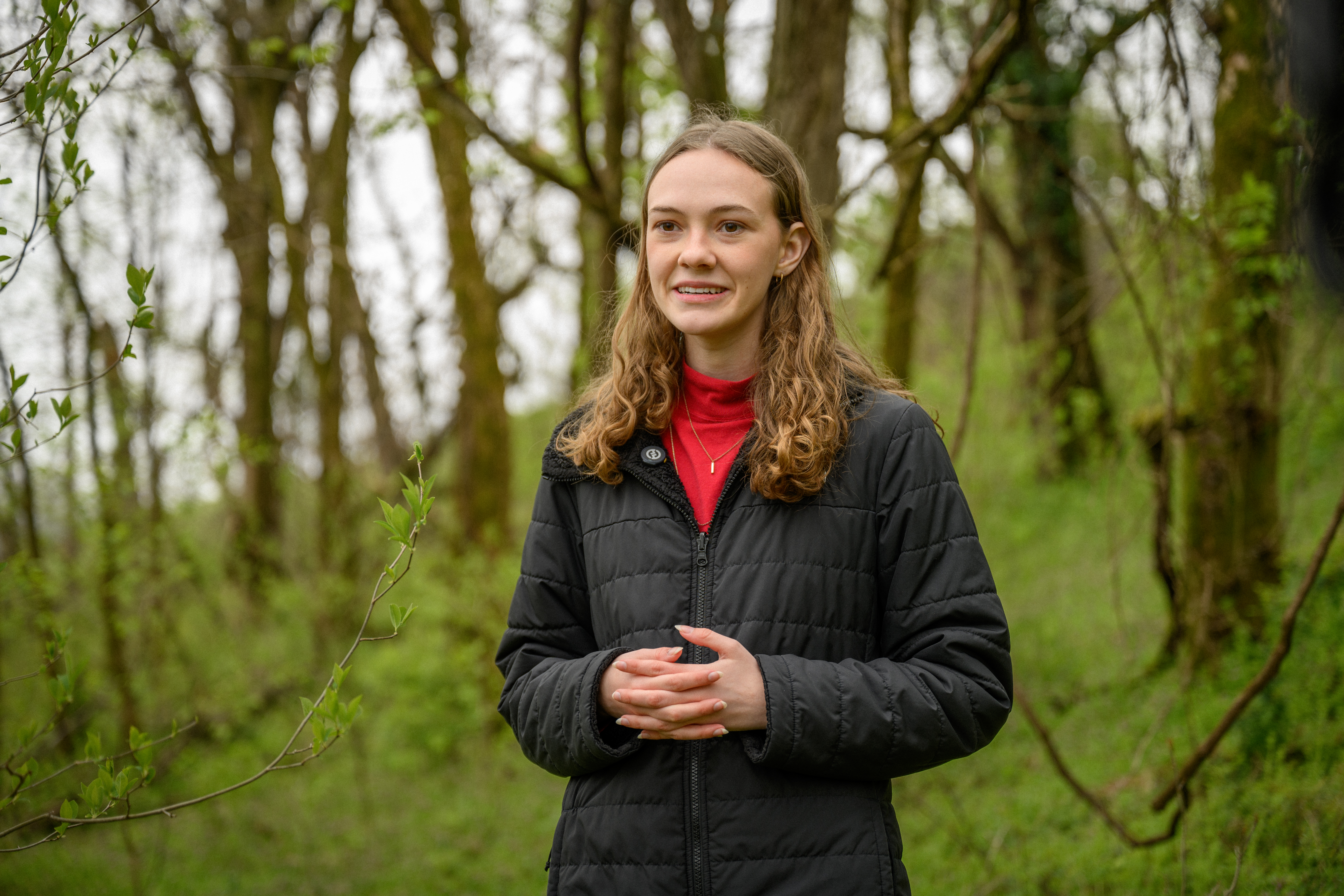 A young woman in a black coat smiles and speaks with her hands while being interviewed in a forest.