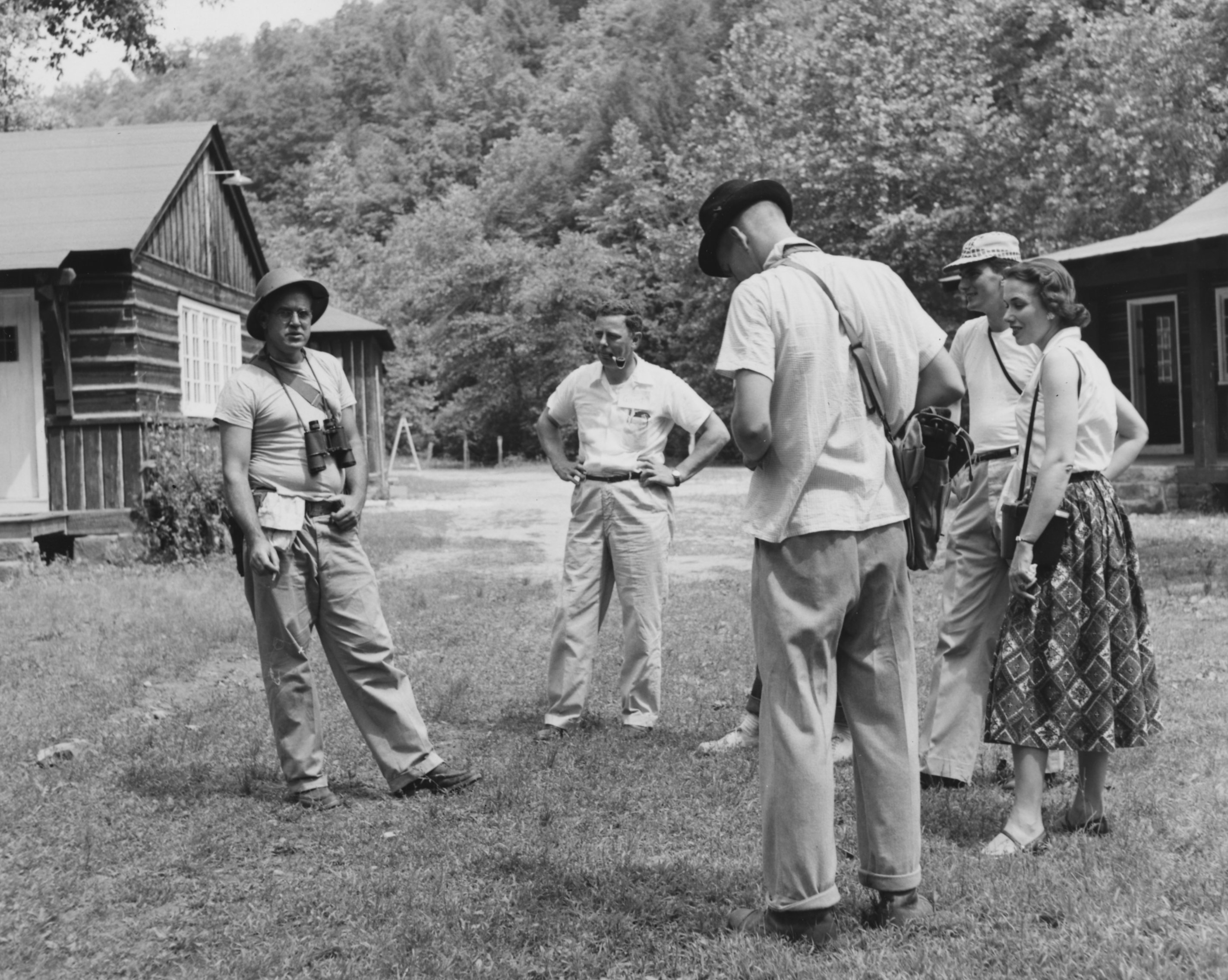 In a black-and-white photo, a group of men and women stand and talk in a camp.