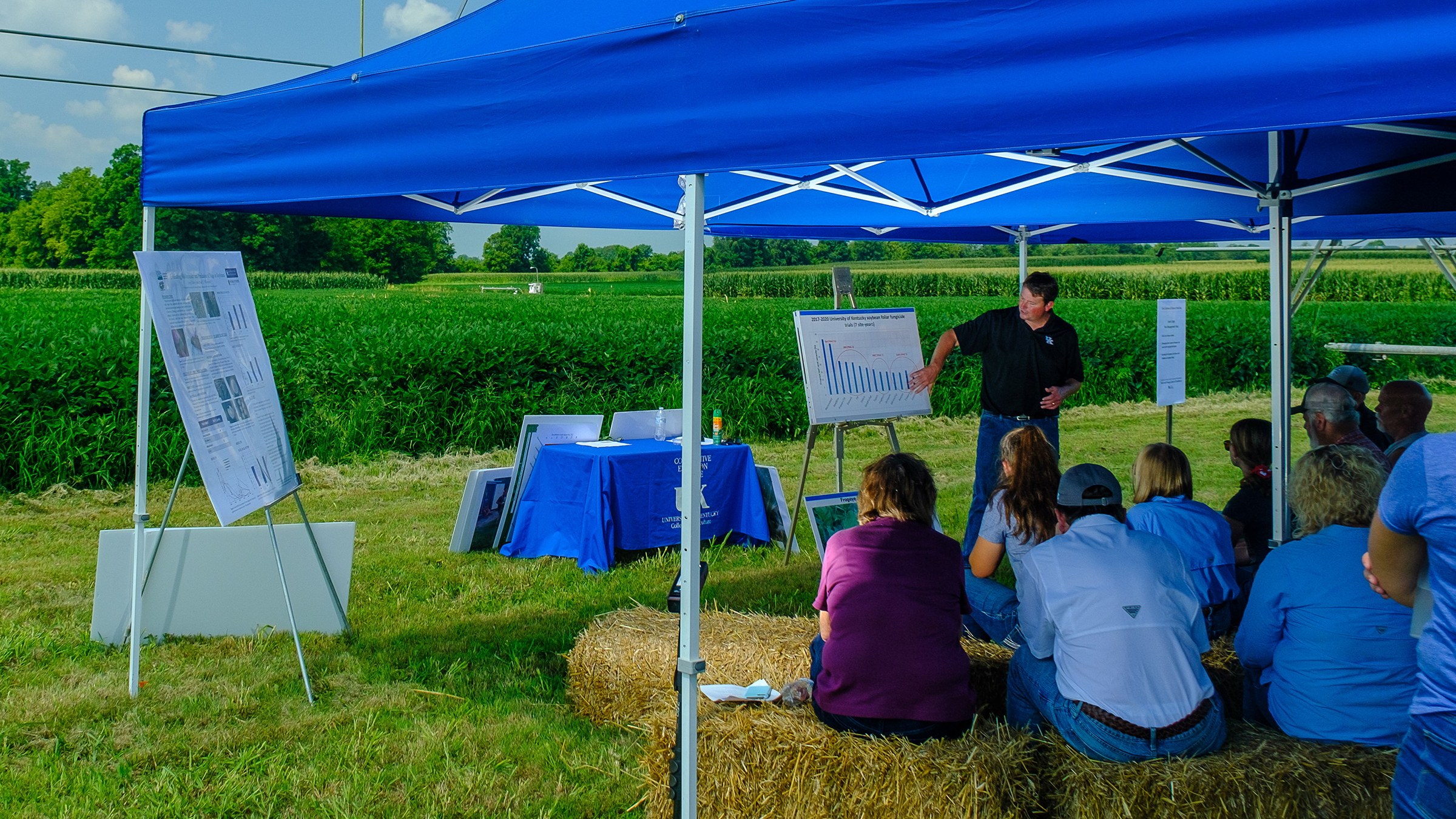 A man gives a presentation in front of a field, while listeners sit on hay bales under a blue tent.