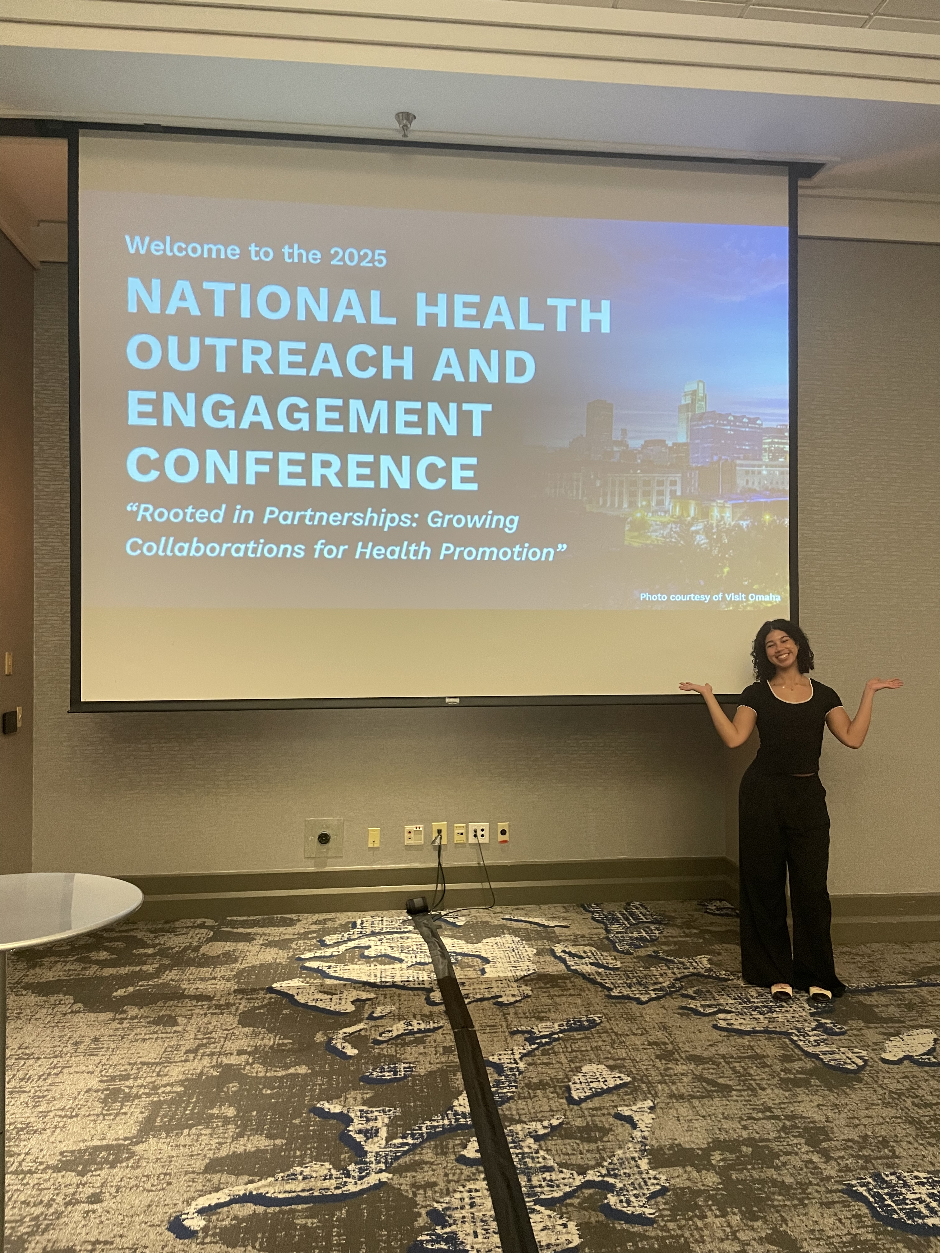 A young woman smiles and gestures proudly in front of a powerpoint screen, which reads Welcome to the 2025 National Health Outreach and Engagement Conference."