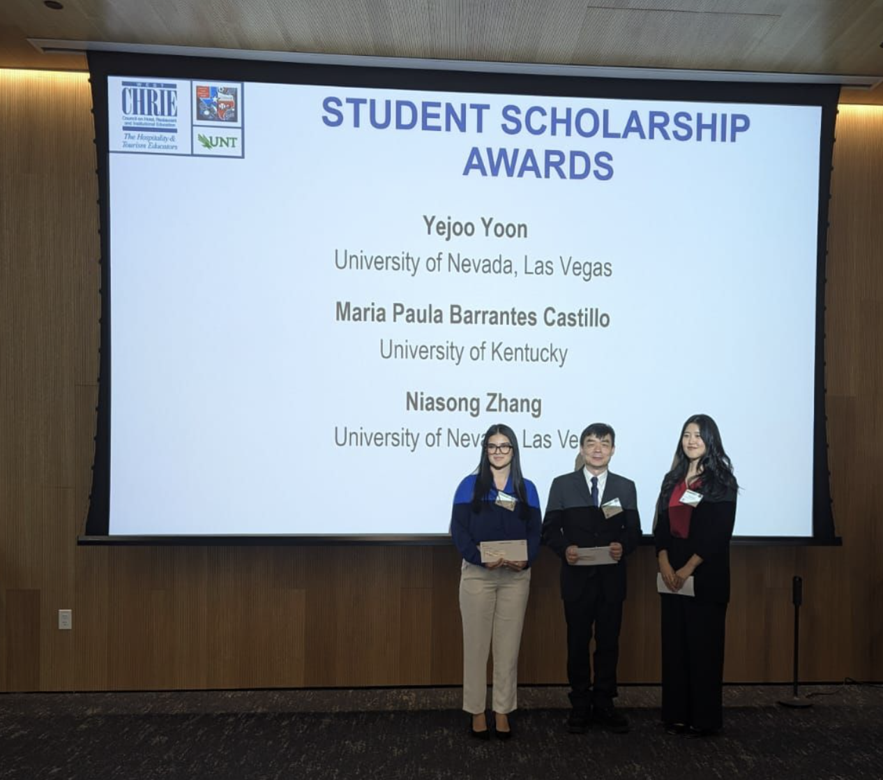 Three young people stand in front of a presentation slide that announces them as scholarship winners.