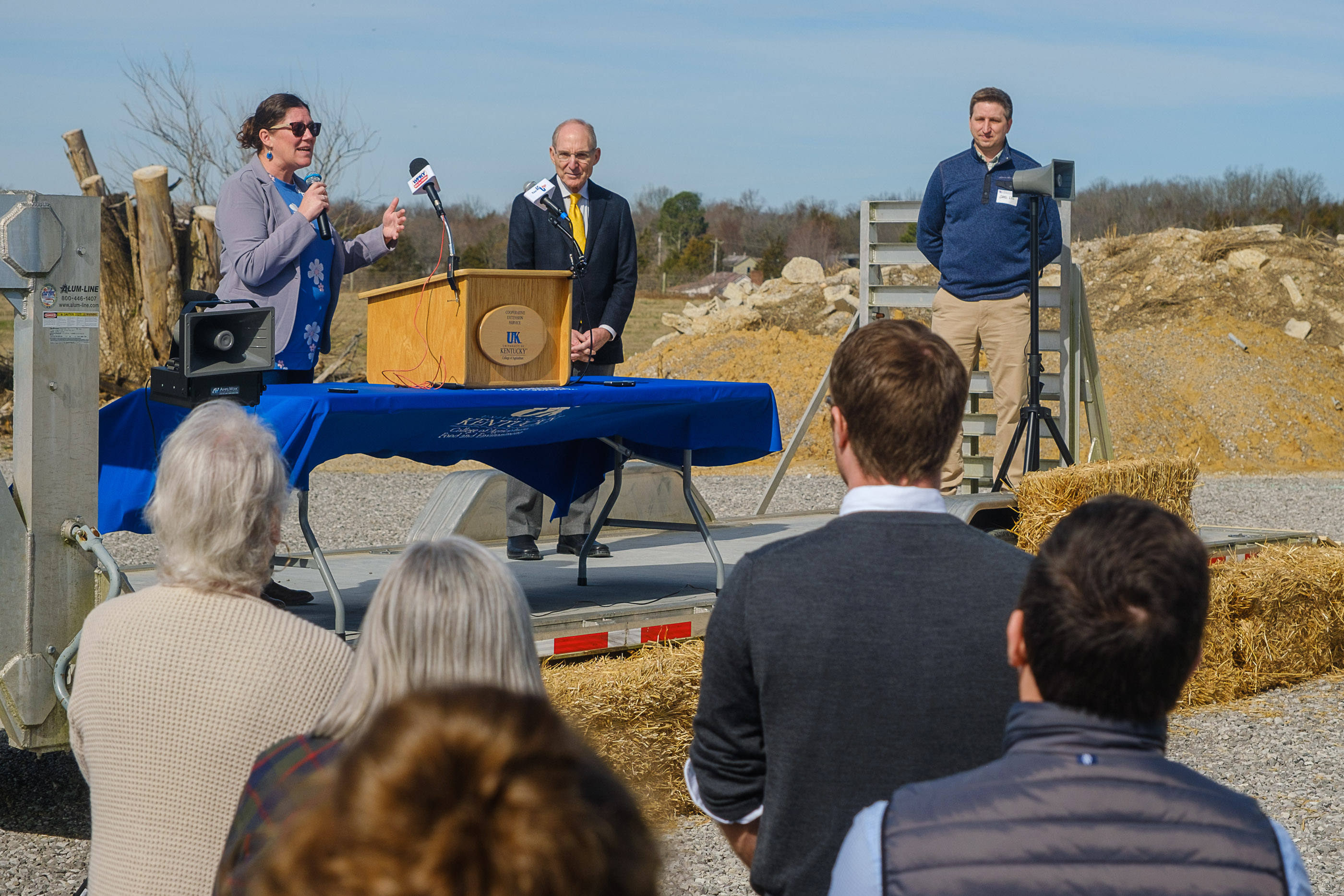 A woman speaks at a makeshift podium on a stage outdoors. Two men stand on the stage beside her.