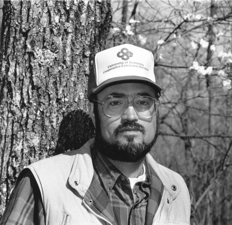In a black-and-white photo, a man is in the forest, wearing a hat that says University of Kentucky Cooperative Extension Service. 