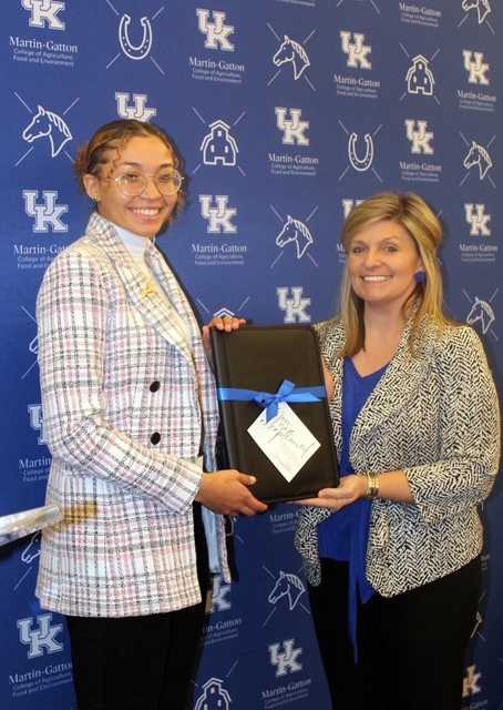 Two women pose for a photo, holding a gift in front of a backdrop that says UK Martin-Gatton College of Agriculture, Food and Environment.