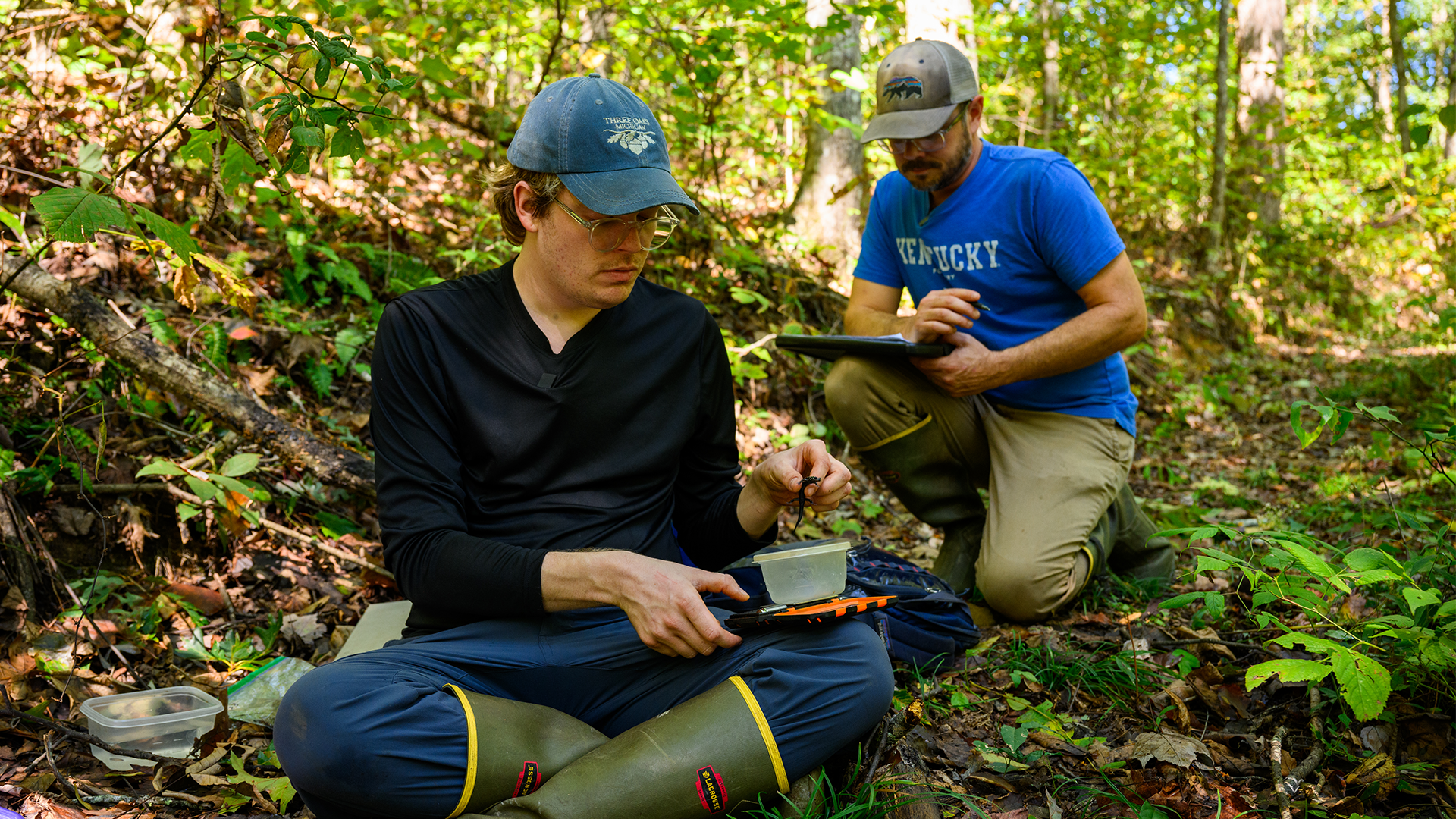 Two men sit on a forest trail, using scientific equipment to study a small black salamander.