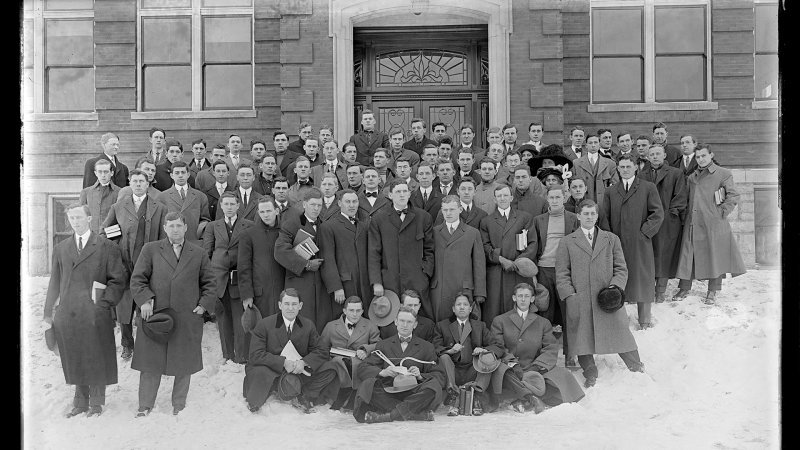 A black-and-white film photo shows a large group of University of Kentucky students posing in front of Scovell Hall in the snow. 