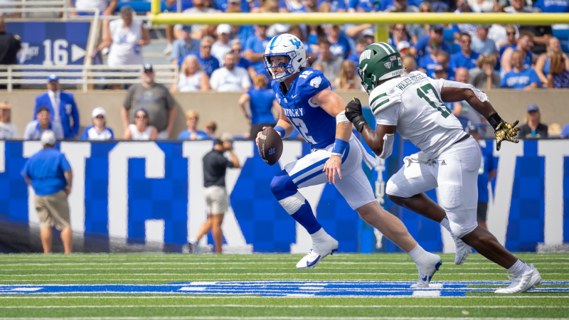 A football player in a blue-and-white uniform holds a football and runs around an opposing player.