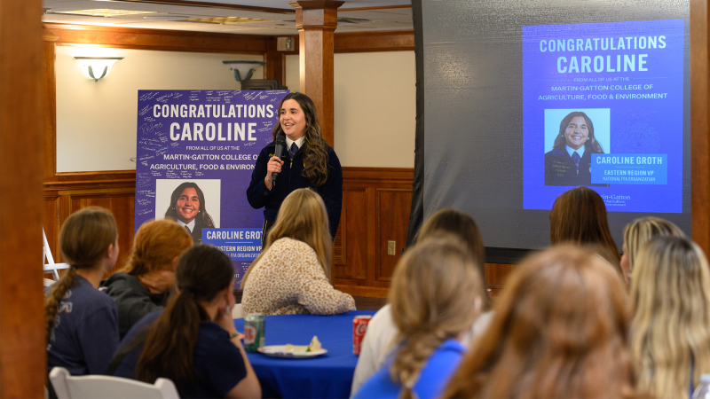 A young woman in a navy FFA jacket speaks in front of a room of people. The screen shows a photo of her and "Congratulations Caroline"