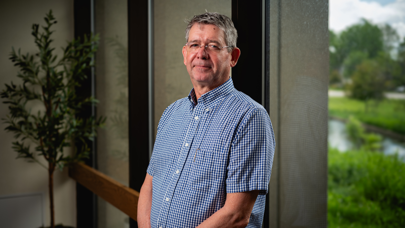 A man in a blue short-sleeve button-up poses for a photo inside an office space with a window behind him.