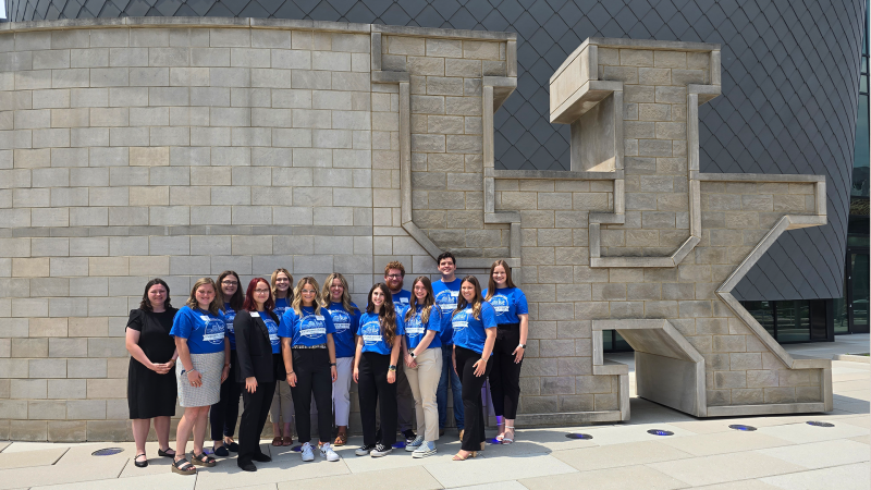 A group of college students and staff pose for a photo next to a large concrete UK logo.