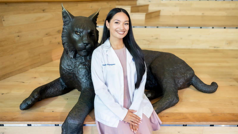 A young woman wearing a light pink dress and a white dietitian's coat sits beside a bronze sculpture of a wildcat, the University of Kentucky's mascot.