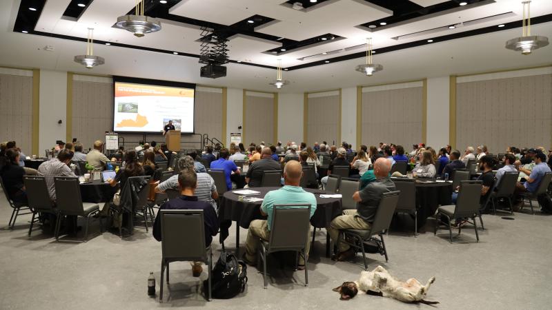 Ballroom full of round tables with folks listening to a speaker at the screen