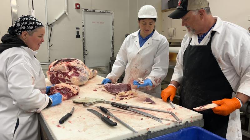 Three people stand around a table as they carve a beef carcass into cuts of meat. 