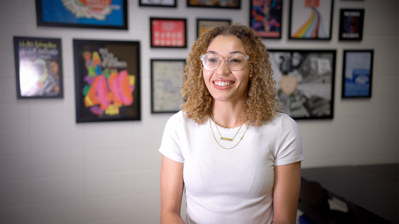 A woman wearing glasses and a white t-shirt smiles. Behind her is a gallery wall of framed photos.