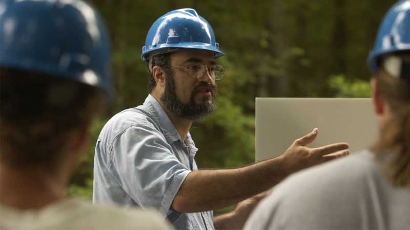 A man wearing a blue hard hat speaks and demonstrates with his hands to some listeners, also wearing blue hard hats.