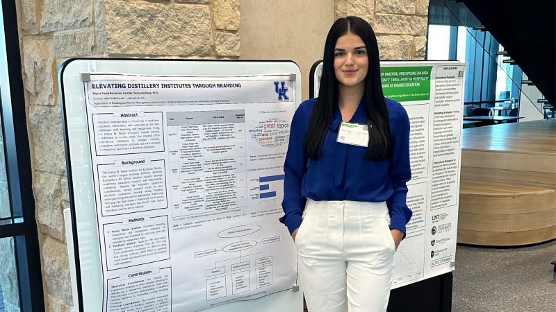 A young woman wearing white pants and a blue top poses next to her research poster. 