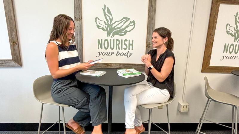 Two women sit at a small round table in front of a food poster with nutrition books in front of them.