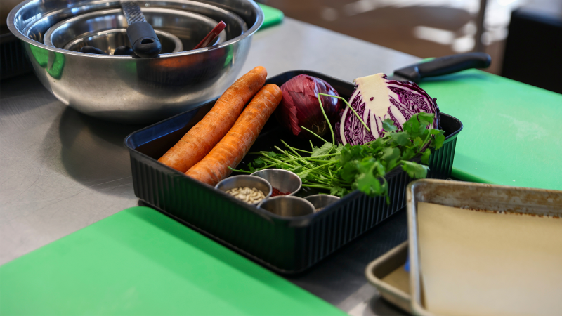 A kitchen counter holds several pieces of cooking equipment and ingredients, such as onion and carrots.