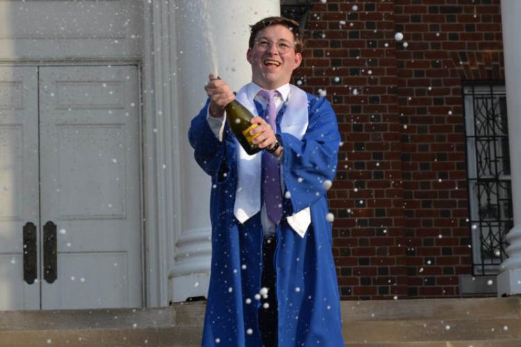 Zachary Chaney in graduation cap and gown popping a bottle of champagne on the steps of UK's Memorial Hall.