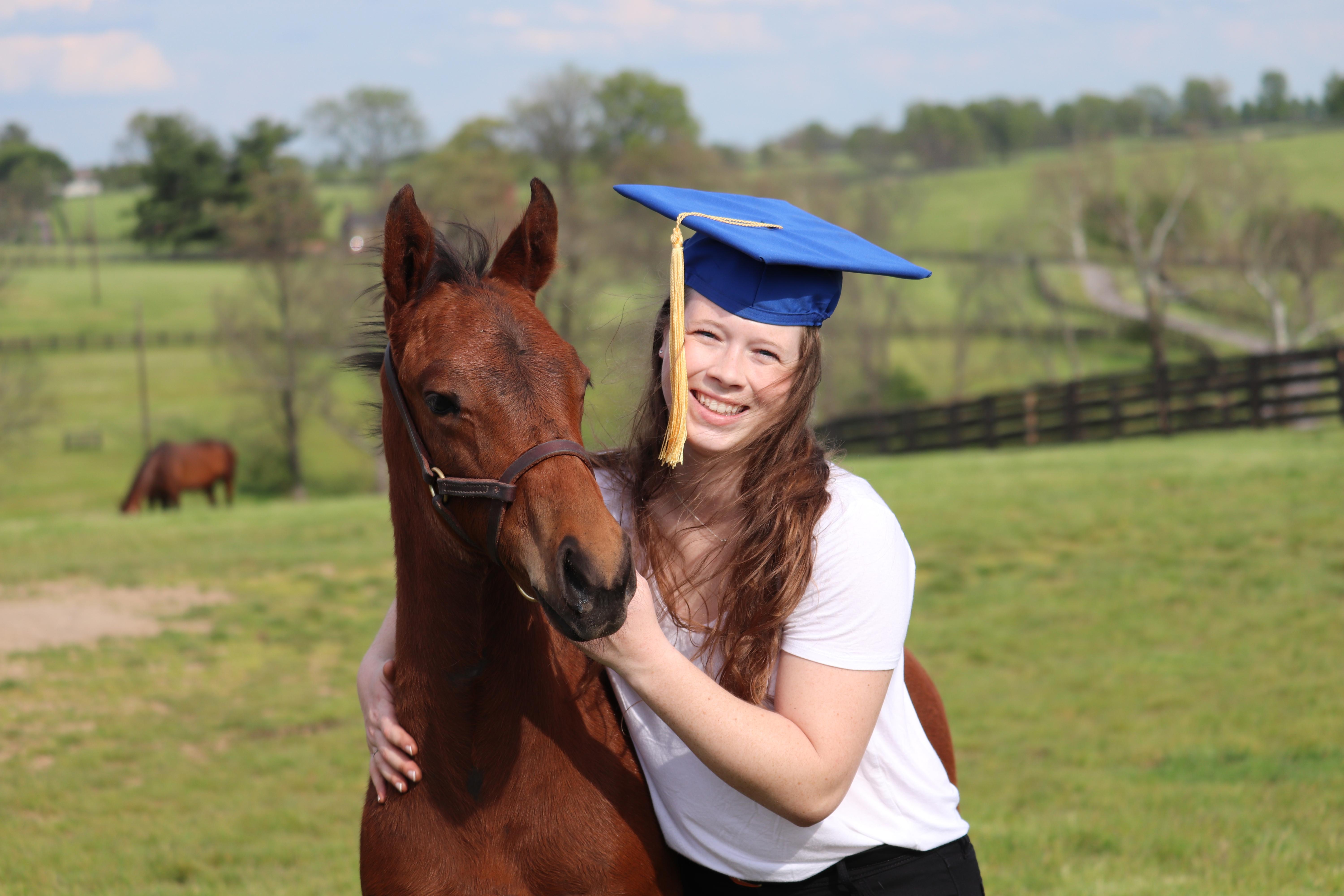 Hannah Moriarty in graduation cap, standing next to horse. 